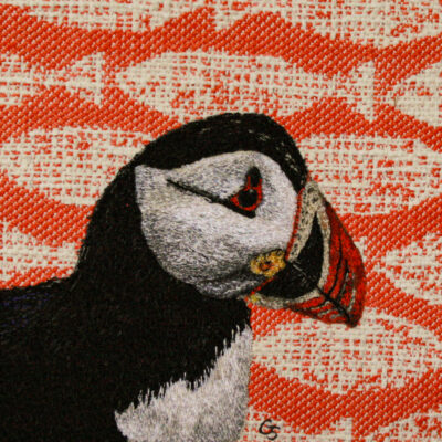 An original free-motion, machine embroidered portrait of a puffin, against a vibrant background of printed white fish on orange.
