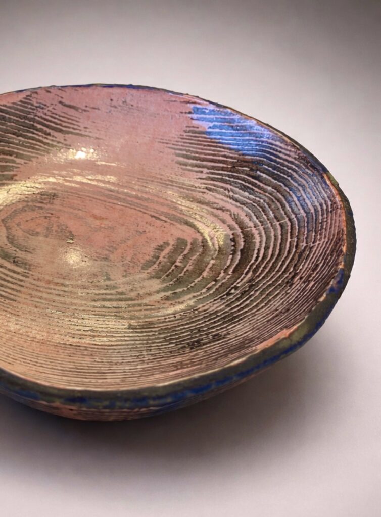 Handmade ceramic bowl with wood grain pattern transferred from a wooden former using slip and underglaze, featuring layered pink, and dark tones, photographed on a neutral exhibition backdrop